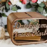 Drewniana skrzynka na pieniądze z wesela Ani i Karol. W tle kwiaty i świeca.