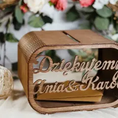 Drewniana skrzynka na pieniądze z wesela Ani i Karol. W tle kwiaty i świeca.