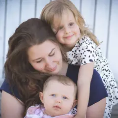 Uśmiechnięta mama tuli swoje dwie córeczki. Starsza dziewczynka obejmuje ją od tyłu, a malutka siedzi na jej rękach. Widać, jaka jest mama kochana.