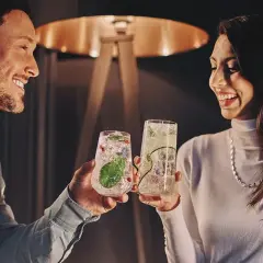 Uśmiechnięta para wznosi toasty drinkami. Idealne pomysły na randki w domowym zaciszu.