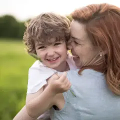 Uśmiechnięty chłopiec przytula się do mamy na tle zielonej łąki. Widać, że to dobra mama, pełna miłości i radości.