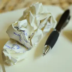 Zgniecione kartki z notatkami i długopis na otwartym notatniku. Proces tworzenia tekstu, jak pisać z osobą towarzyszącą, bywa pełen prób i błędów.