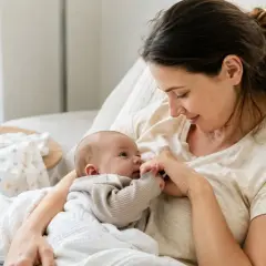 Delikatny uścisk dłoni dziecka i matki, symbolizujący budowanie więzi. Na stoliku butelka z mlekiem, obok otulacz.