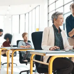 Mężczyzna udziela kobiecie konstruktywnej krytyki przy komputerze. W tle pracują inni ludzie.
