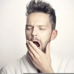 Mężczyzna w szlafroku ziewa, zakrywając usta dłonią. To może oznaczać ziewanie podczas rozmowy, gdy brakuje mu snu lub nudzi się.