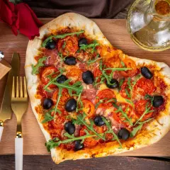 Serce z pizzy z pomidorami, oliwkami i rukolą, przygotowane na romantyczną kolację dla żony. Obok złote sztućce i młynek do pieprzu.