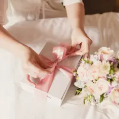 Ręce zawiązują różową wstążkę na pudełku prezentowym. Obok leży bukiet pastelowych kwiatów. To piękny dzień chrzestnej.