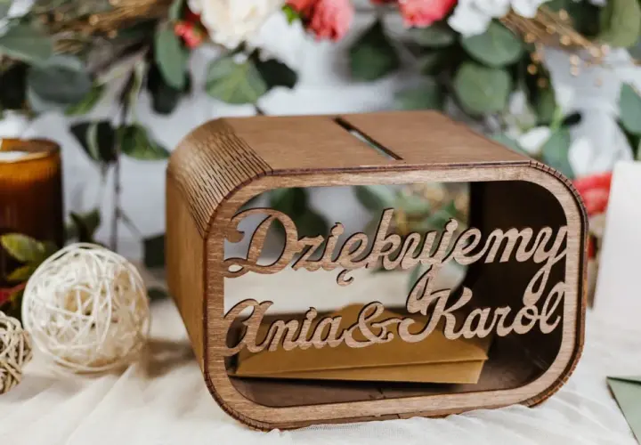 Drewniana skrzynka na pieniądze z wesela Ani i Karol. W tle kwiaty i świeca.