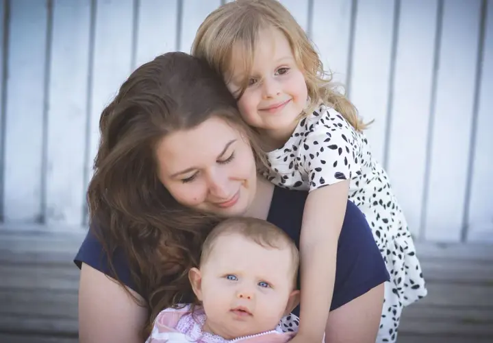 Uśmiechnięta mama tuli swoje dwie córeczki. Starsza dziewczynka obejmuje ją od tyłu, a malutka siedzi na jej rękach. Widać, jaka jest mama kochana.
