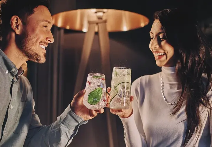 Uśmiechnięta para wznosi toasty drinkami. Idealne pomysły na randki w domowym zaciszu.