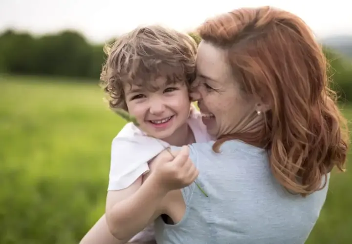 Uśmiechnięty chłopiec przytula się do mamy na tle zielonej łąki. Widać, że to dobra mama, pełna miłości i radości.
