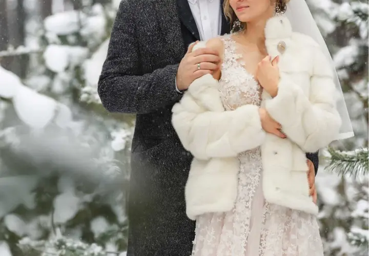 Para młoda w zimowej scenerii. Ona w białym futerku i sukni ślubnej, on w eleganckim płaszczu. Ich ślub zimą jest pełen uroku.