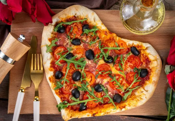 Serce z pizzy z pomidorami, oliwkami i rukolą, przygotowane na romantyczną kolację dla żony. Obok złote sztućce i młynek do pieprzu.