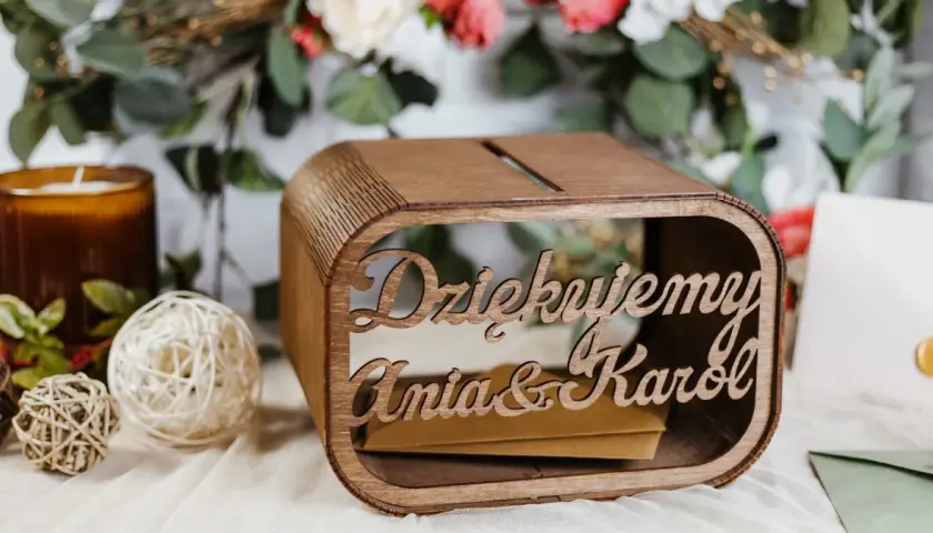 Drewniana skrzynka na pieniądze z wesela Ani i Karol. W tle kwiaty i świeca.