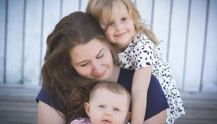 Uśmiechnięta mama tuli swoje dwie córeczki. Starsza dziewczynka obejmuje ją od tyłu, a malutka siedzi na jej rękach. Widać, jaka jest mama kochana.