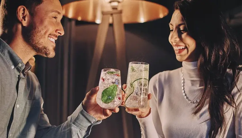 Uśmiechnięta para wznosi toasty drinkami. Idealne pomysły na randki w domowym zaciszu.