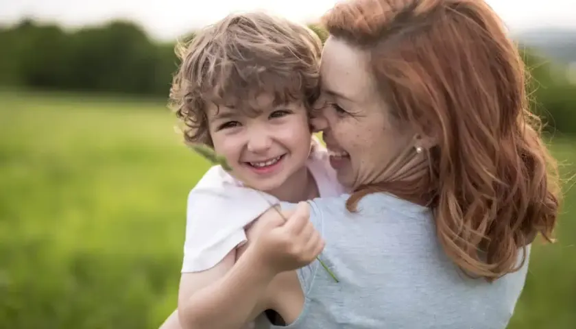 Uśmiechnięty chłopiec przytula się do mamy na tle zielonej łąki. Widać, że to dobra mama, pełna miłości i radości.