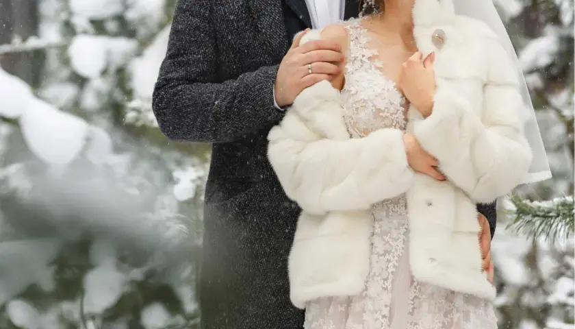 Para młoda w zimowej scenerii. Ona w białym futerku i sukni ślubnej, on w eleganckim płaszczu. Ich ślub zimą jest pełen uroku.