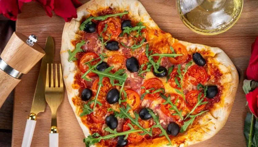 Serce z pizzy z pomidorami, oliwkami i rukolą, przygotowane na romantyczną kolację dla żony. Obok złote sztućce i młynek do pieprzu.