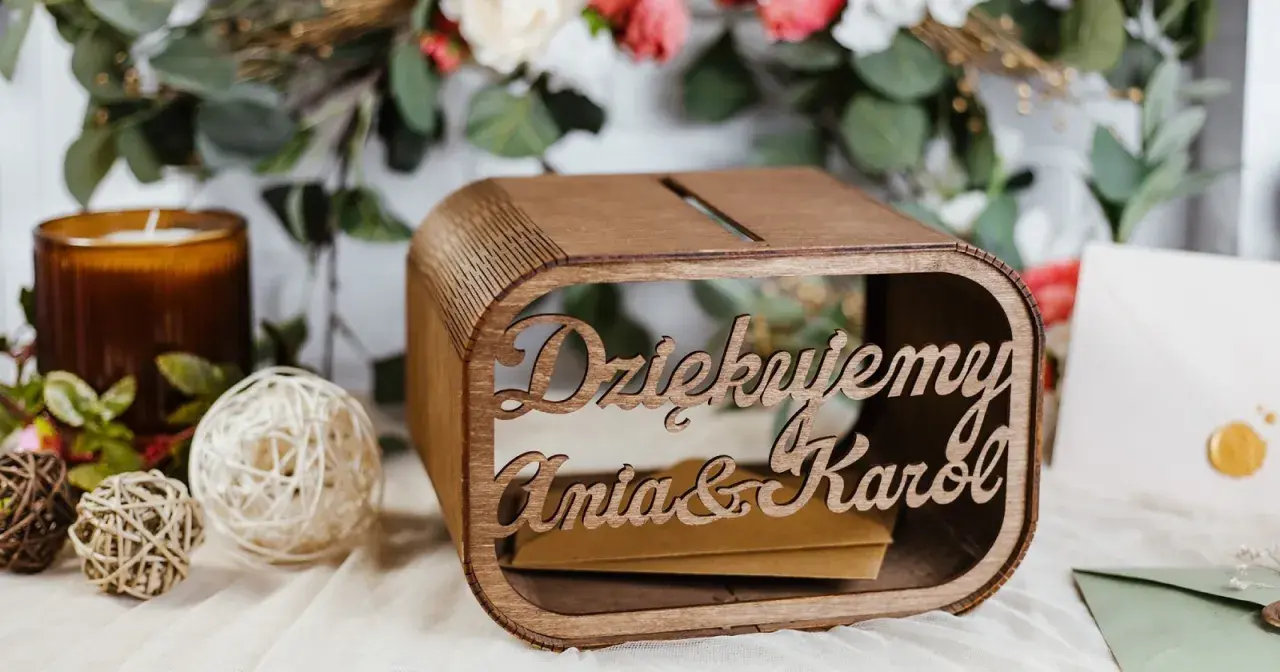 Drewniana skrzynka na pieniądze z wesela Ani i Karol. W tle kwiaty i świeca.