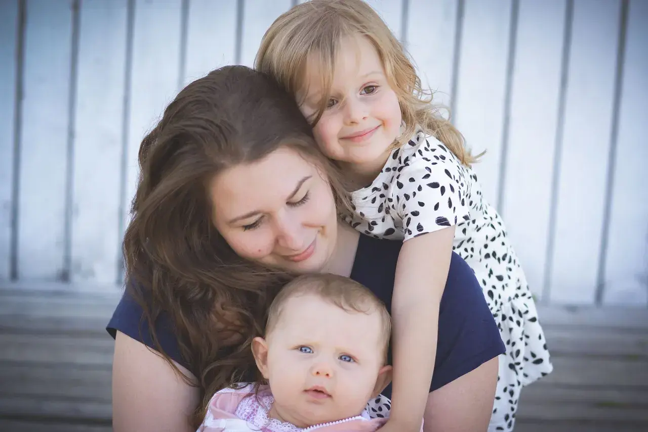 Uśmiechnięta mama tuli swoje dwie córeczki. Starsza dziewczynka obejmuje ją od tyłu, a malutka siedzi na jej rękach. Widać, jaka jest mama kochana.