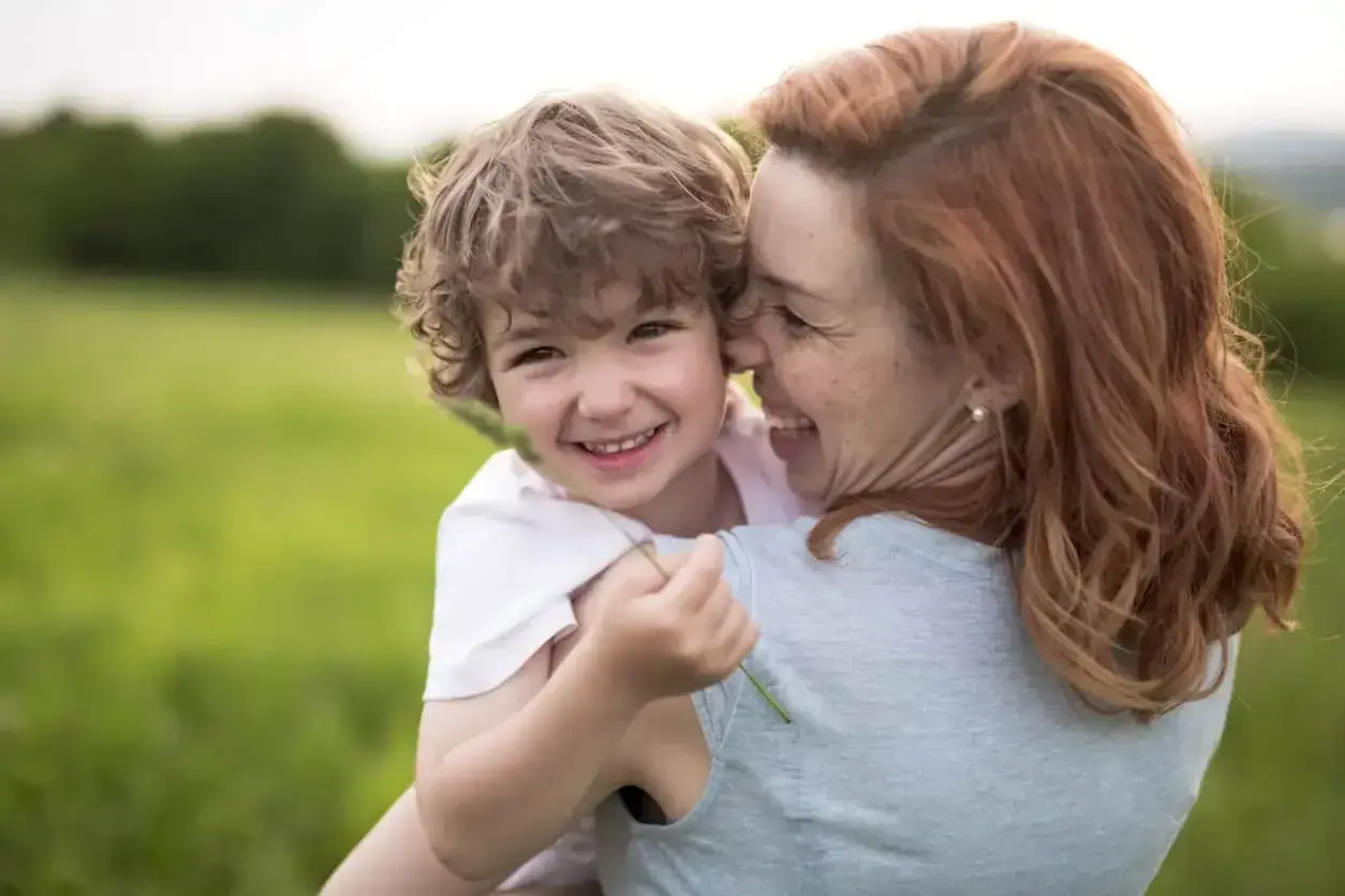 Uśmiechnięty chłopiec przytula się do mamy na tle zielonej łąki. Widać, że to dobra mama, pełna miłości i radości.
