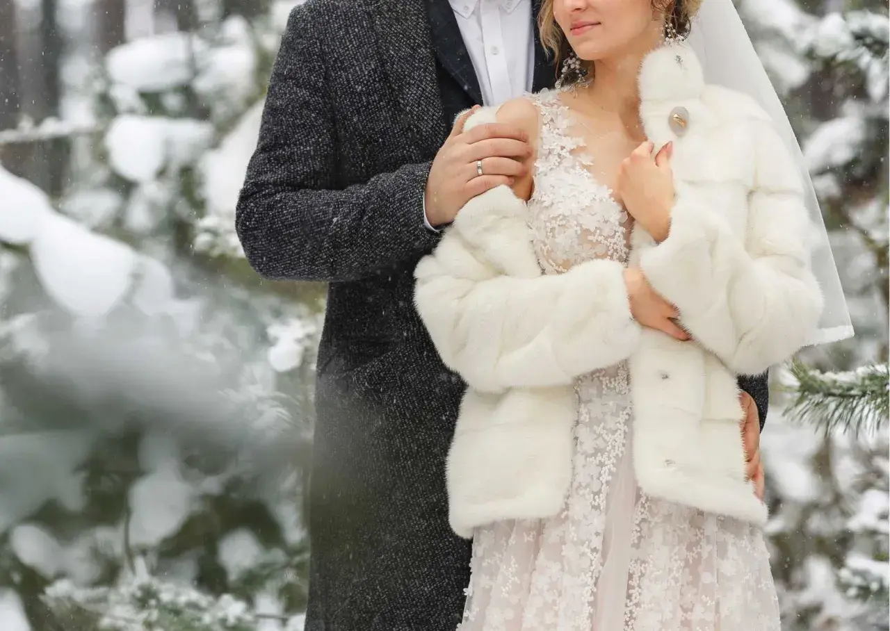 Para młoda w zimowej scenerii. Ona w białym futerku i sukni ślubnej, on w eleganckim płaszczu. Ich ślub zimą jest pełen uroku.