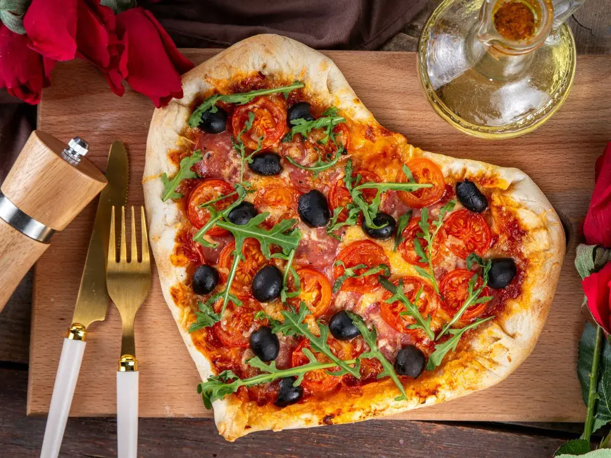 Serce z pizzy z pomidorami, oliwkami i rukolą, przygotowane na romantyczną kolację dla żony. Obok złote sztućce i młynek do pieprzu.