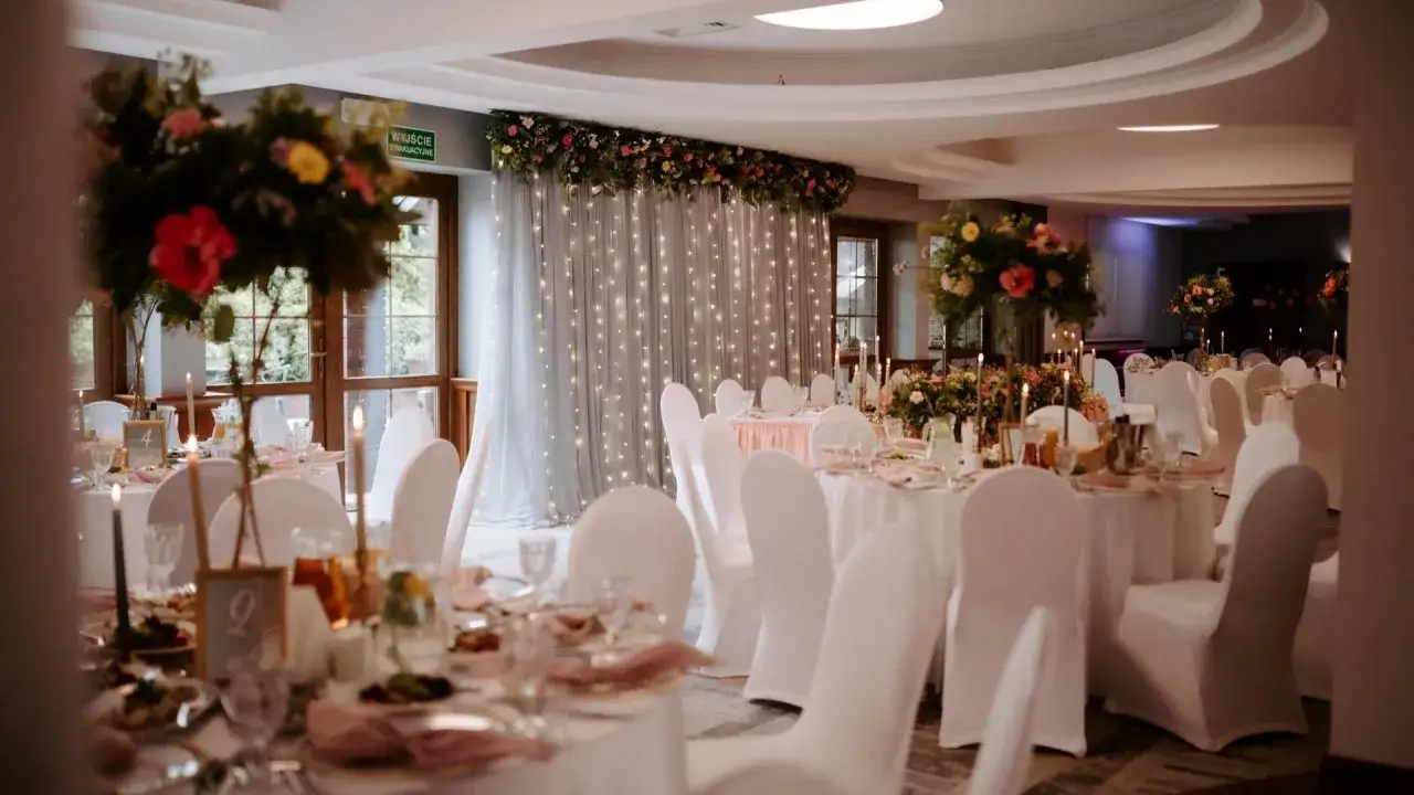 Elegancka sala weselna z białymi pokrowcami na krzesłach i kwiecistymi dekoracjami. Idealne miejsce, by napisać maila o salę weselną i zaplanować wymarzone przyjęcie.