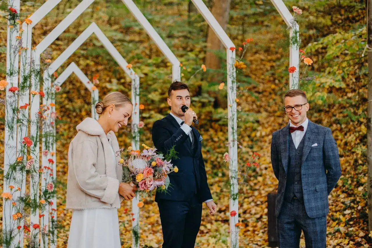 Piękny ślub we wrześniu. Panna młoda z bukietem, pan młody m&oacute;wi do mikrofonu, a świadek uśmiecha się w kratowanym garniturze.