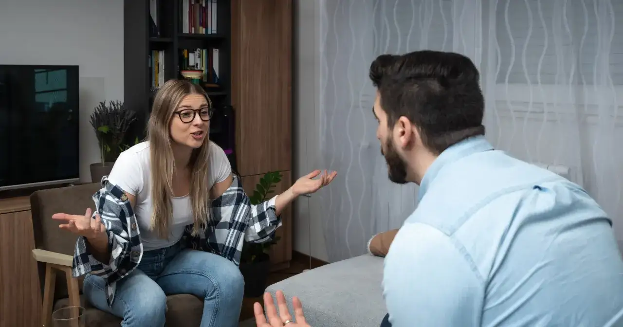 Kłótnia małżeńska w salonie. Kobieta w okularach gestykuluje, mężczyzna siedzi tyłem.