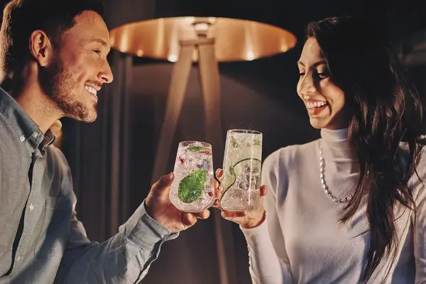 Uśmiechnięta para wznosi toasty drinkami. Idealne pomysły na randki w domowym zaciszu.